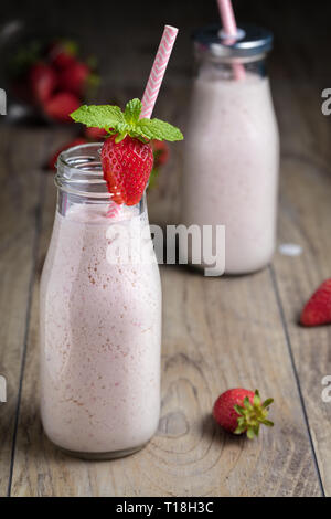 Un sano frullato fragole, milk shake, nelle tradizionali bottiglie con bacche sparse e paglia sopra uno sfondo di legno. Composizione verticale Foto Stock