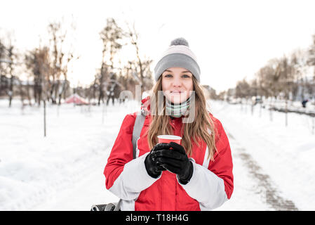 Una ragazza studente in inverno in città sullo sfondo di neve e strada, nella sua mano trattiene una tazza di caffè, tè, si riscalda in una fredda giornata invernale. Emozioni di Foto Stock