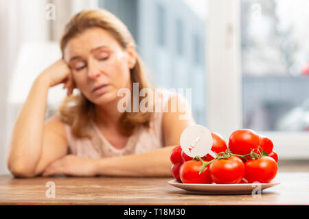 Casalinga seduta a tavola e guardando i pomodori ma avente allergia Foto Stock