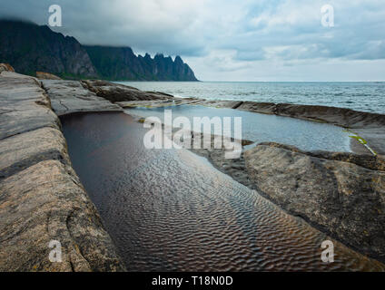La spiaggia sassosa con bagni di marea a Ersfjord, Senja, Norvegia. Estate giorno polare notte costa. I denti di drago di roccia nel lontano. Foto Stock