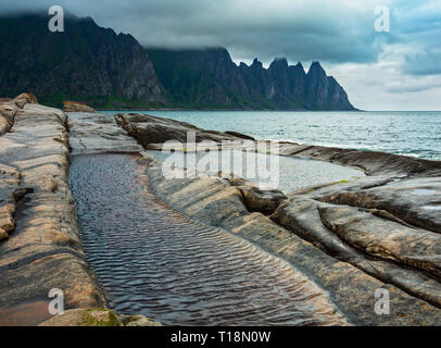 La spiaggia sassosa con bagni di marea a Ersfjord, Senja, Norvegia. Estate giorno polare notte costa. I denti di drago di roccia nel lontano. Foto Stock