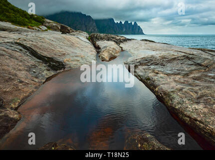 La spiaggia sassosa con bagni di marea a Ersfjord, Senja, Norvegia. Estate giorno polare notte costa. I denti di drago di roccia nel lontano. Foto Stock