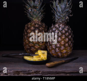 Ananas a fette sulla vecchia scrivania rustico con coltello e con due ananas dietro. Sfondo nero. Foto Stock