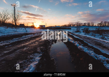 Paese su strada sterrata con pozzanghere fangose e la neve che si scioglie in primavera al tramonto Foto Stock