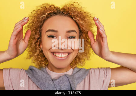 E attraente ragazza positivo sta cercando di fotocamera. Lei tiene le mani vicino alla sua testa. Isolato su sfondo giallo Foto Stock