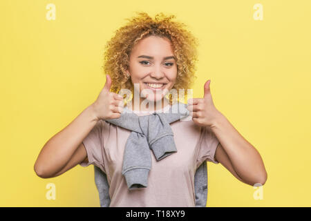 Bello e attraente ragazza sta tenendo in mano il suo grande pollice in alto su entrambi le mani e sorridente. Lei è alla ricerca sulla fotocamera. Isolato su sfondo giallo Foto Stock