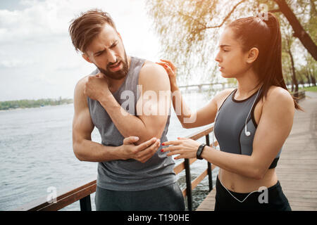 Il ragazzo è in piedi con una ragazza su un ponte. Egli ha un dolore nel suo gomito. La sua ragazza sta tenendo in mano la sua mano sulla sua spalla e preoccuparsi di lui Foto Stock