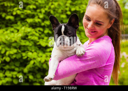 Amante degli animali, animali domestici di piccola taglia, cani concetto di divertimento. Sorridenti donna che indossa indumenti sportivi azienda piccola carino bulldog francese nel parco al di fuori Foto Stock