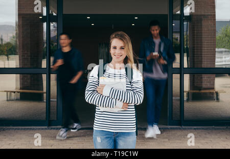 Fiducioso studentessa permanente al campus universitario con altri studenti a piedi in background. Giovane donna in piedi in college con studenti visitatore occasionale Foto Stock