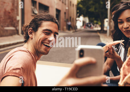 Sorridente giovane uomo prendendo selfie con amici avente la pizza. Amici prendendo selfie mentre mangiare la pizza seduti all'aperto sulla strada della citta'. Foto Stock