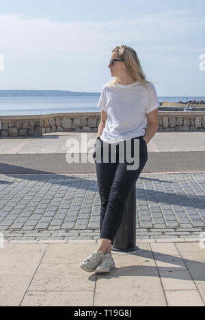 Giovane sportivo casual vestito donna bionda con occhiali da sole in t-shirt bianco si siede vicino alla strada con ocean dietro a Mallorca, Spagna. Foto Stock