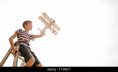 Ragazzo seduto sulla cima di una scala con aeroplano giocattolo contro la luce diretta del sole. Ragazzo immagina di essere pilota, volare un aereo superiore. Foto Stock