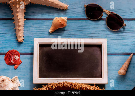 Da sopra la lavagna tra gli occhiali da sole e stelle di mare con conchiglie blu su sfondo di legno Foto Stock