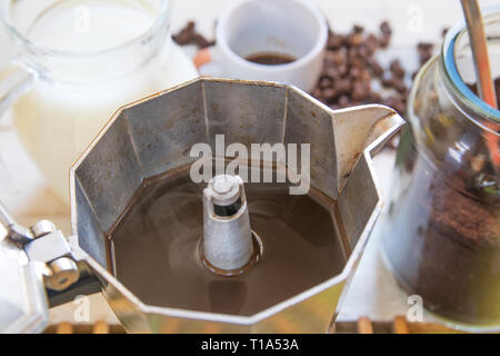 Teiera con alcuni caffè nero su un tavolo di legno accanto a chicchi di caffè, latte e una coppa in una cucina rustica. Foto Stock