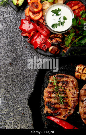 Bistecche alla griglia in ghisa padella per grigliare su sfondo nero con una varietà di verdure grigliate. Carne alla brace. Vista dall'alto. Copia dello spazio. Foto Stock