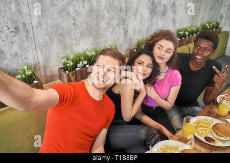 Ragazze e giovani uomini seduti in cafe e facendo selfie. Gruppo di gente allegra colorata in t shirts da tavolo in legno con fast food, hamburger e il succo nei bicchieri. Amici in posa sorridente. Foto Stock