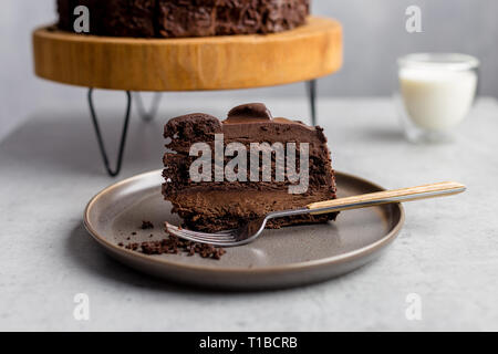 Gustoso pezzo di torta al cioccolato su lastra grigia con forcella, con tutta la torta di cacao su supporto in legno, bicchiere di latte vicino su sfondo grigio. Vista laterale. Concep Foto Stock