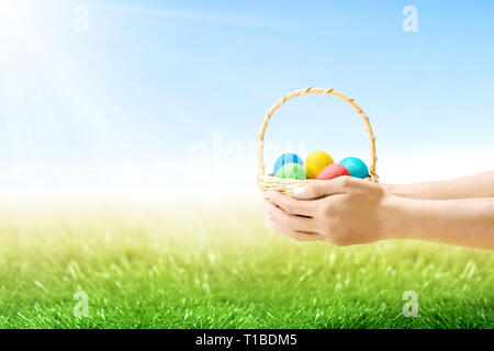 Le mani umane holding cesto in legno con colorate uova di pasqua sul campo di erba con la luce del sole e cielo blu sullo sfondo. Buona Pasqua Foto Stock