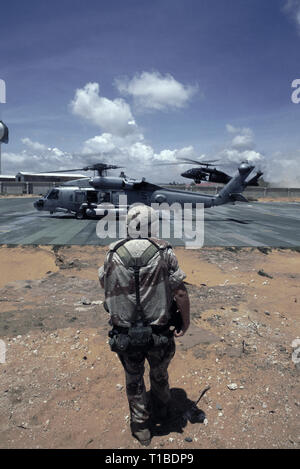 Il 29 ottobre 1993 A U.S. Soldato dell'esercito si erge in primo piano come un US Navy Sikorsky SH-60 Seahawk elicottero dall'USS Abraham Lincoln si prepara per il decollo dalla heli-pad a UNOSOM HQ a Mogadiscio, in Somalia. In background, un esercito americano UH-60 Black Hawk elicottero è attorno alla terra. Foto Stock