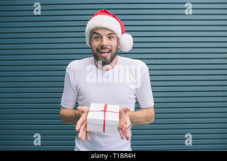 Eccitato uomo barbuto è in piedi e tenendo presente le mani. Egli è stupito. L uomo indossa cappello di Natale. Isolato su striped e sfondo blu. Foto Stock
