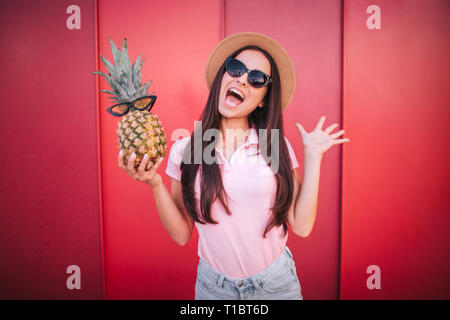 Felice ed emozionato brunette sorge e ananas tiene in una mano. Esso dispone di occhiali da sole. La ragazza lo indossa come bene. Ella è stupito. Isolato su rosso e Foto Stock