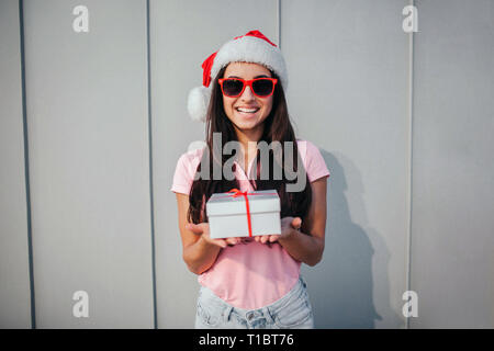 Allegro ragazza nel cappello di Natale si erge e tiene presenti nelle mani. Lei si guarda sulla fotocamera e sorrisi. Isolato su striped e sfondo bianco. Foto Stock