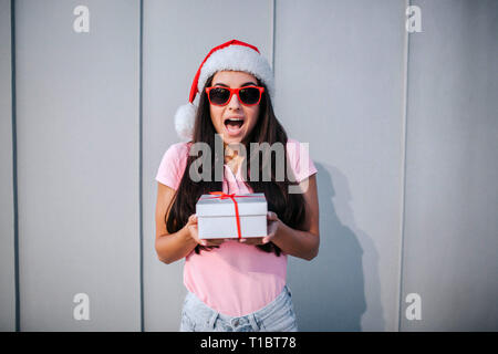 Stupiti ed entusiasti di ragazza si erge e detiene il riquadro bianco con striscia rossa. Lei si guarda sulla telecamera attraverso occhiali da sole. Isolato su striped e sfondo bianco. Foto Stock