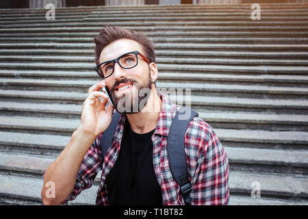 Bella giovane uomo seduto sulle scale e guardare in alto. Egli parla al telefono. Ragazzo indossa occhiali. Egli è in attesa. Giovane uomo è felice. Foto Stock