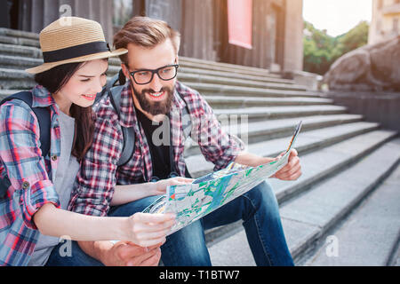 Giovane uomo e donna sedersi sui passi al di fuori. Essi detengono mappa insieme. I turisti sorriso. Essi guardano alla mappa. I viaggiatori hanno il resto. Foto Stock