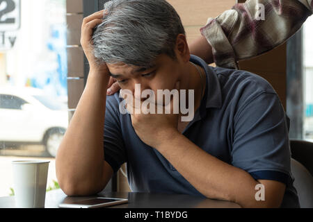 Di mezza età uomo asiatico 40 anni, ha sottolineato e stanco, sono seduti in un ristorante fast food e avere amici in piedi dietro a incoraggiare. Concetto di Foto Stock