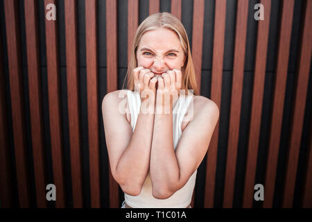 Arrabbiato ragazza è in piedi a parete e alla ricerca sulla fotocamera. Ella è molto turbato. Giovane donna sta tenendo le dita sulla bocca. Isolato su sfondo a strisce. Foto Stock