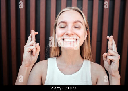 Felice e positivo ragazza bionda è in piedi e tenere gli occhi chiusi. Lei è sorridente. Giovane donna sta attraversando la sua fingr su entrambe le mani. Isolato su Foto Stock