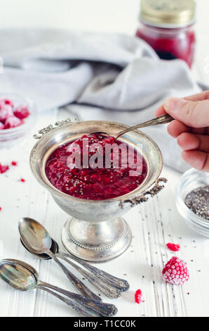 In casa confettura di lamponi con semi di chia su sfondo bianco con womans mano azienda cucchiaio. Una sana idea per colazione pane tostato. Superfoods e sano f Foto Stock