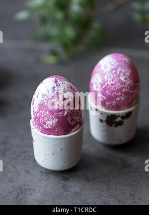 Due multicolore di uova di Pasqua in uovo bianco-cup su sfondo scuro. Concetto di Pasqua gastronomica Foto Stock