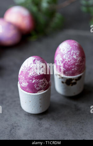 Due colorate uova di Pasqua in uovo bianco-cup su sfondo scuro. Concetto di Pasqua gastronomica Foto Stock