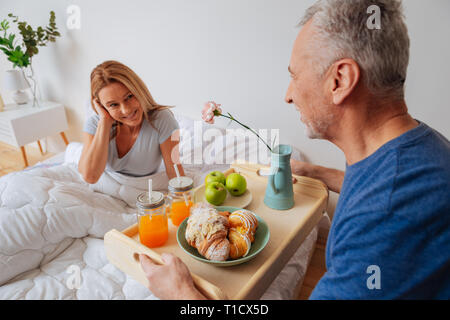 Moglie guardando il marito tenendo il vassoio con la colazione e fiori Foto Stock