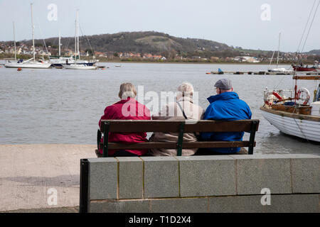 Tre persone anziane seduta su una panchina su Conwy Quayside guardando le barche e yacht nel porto di Conwy Foto Stock