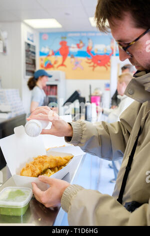 Un uomo mette sale sulla cima del suo pesce e patatine a No1 Fish & Chips Shop a Cromer, North Norfolk, Inghilterra. Foto Stock