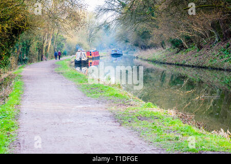 Un giovane a piedi lungo la strada alzaia del Kennet and Avon canal passando houseboats con un haze dai camini delle barche su una luminosa giornata di primavera. Foto Stock