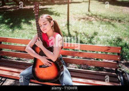 Musicista ragazza è seduta sul banco di lavoro e tenendo la chitarra. Lei è appoggiata su di esso. La ragazza ha chiuso gli occhi e godendo il momento. Foto Stock