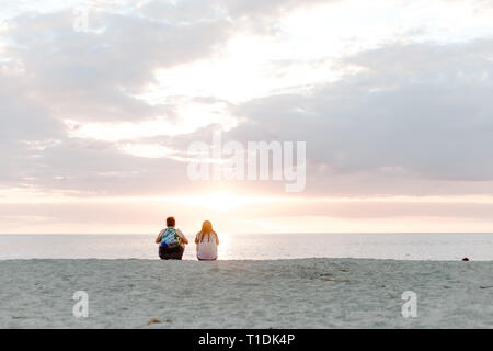 Due persone che guardano il bello e sereno tramonto sull'oceano orizzonte durante la seduta nella sabbia in spiaggia tropicale Foto Stock