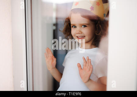 Ritratto di bambina con la corona a casa, guardando fuori della finestra. Shot attraverso il vetro. Foto Stock