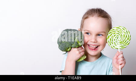 Poco bella ragazza divertirsi con broccoli e lollipop. sceglie cosa mangiare meglio Foto Stock