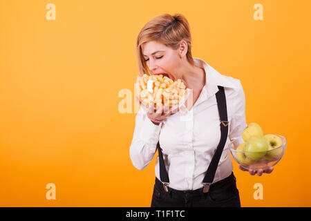 Bella donna bionda con capelli corti prendendo un morso da lei sbuffi di mais su sfondo giallo. Gustosa sbuffi di mais,. Mele fresche. Mele Verdi. Foto Stock