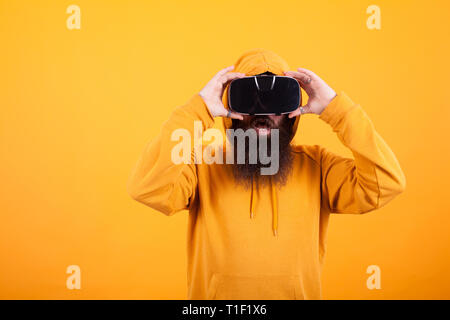 L'uomo con la lunga barba che indossa la realtà virtuale auricolare guardando scioccato su sfondo giallo. Felpa con cappuccio giallo. La tecnologia moderna. Uomo bello. Foto Stock