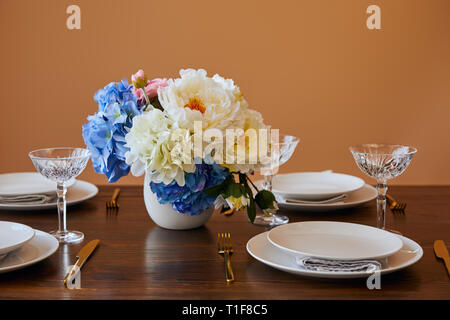 Piastre, golden coltelli e forchette, bicchieri di cristallo e mazzo di fiori in vaso bianco sul tavolo di legno isolata sul beige Foto Stock