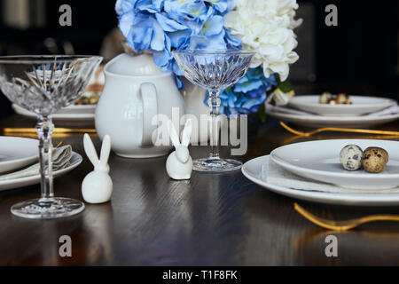 Messa a fuoco selettiva di conigli decorativo vicino a piastre, cristallo e fiori sul tavolo di legno a casa Foto Stock