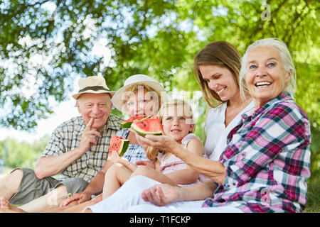 Felice famiglia estesa con il bambino e i nonni mentre mangia il melone nel parco in estate Foto Stock