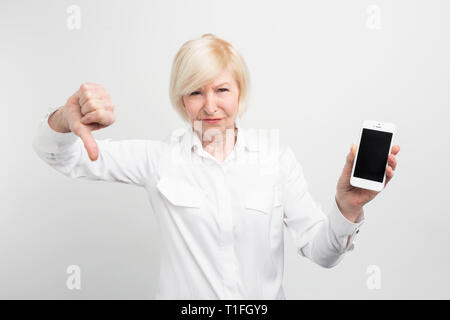 Una foto di donna matura con il nuovo smartphone. Ella ha collaudato e ammesso questo telefono è uno cattivo. Questo è il motivo per cui lei dimostra una grande pollice in basso. Isolato Foto Stock