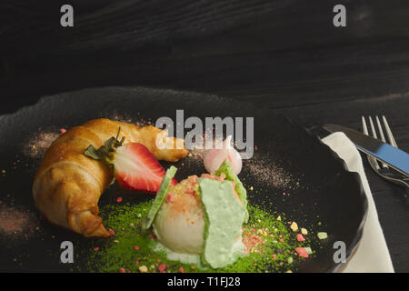 Fetta di strudel di mele con una pallina di gelato alla vaniglia su una piastra nera, su di un legno nero tabella, decorata con meringa e fragole. close-up Foto Stock